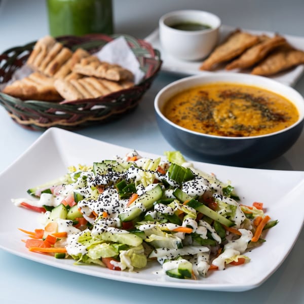 A fresh garden salad with dressing and black sesame seeds, with soup and pita bread in the background.