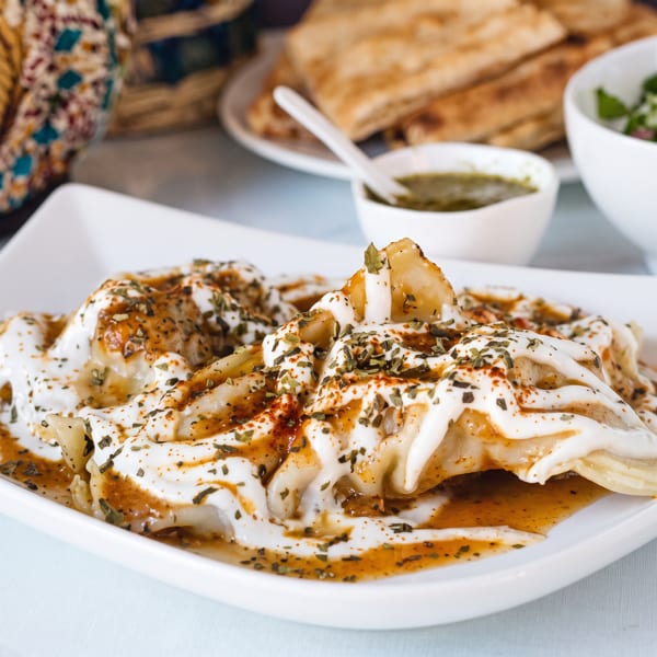Manti dumplings topped with yogurt, spiced oil, and dried herbs, with flatbread and sauce in background.